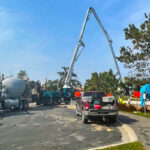 Construction site with cement trucks and a concrete pump in operation. Workers are visible near equipment, with trees and a clear sky in the background.