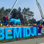 Large blue letters spell "#BEMIDJI" against a backdrop of trees. A crane operates near a blue ox statue. Workers in high-visibility vests are present.