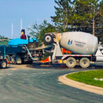 Concrete mixer and pump truck at construction site, with a worker nearby. Trees and a building in the background under a clear blue sky.