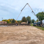 Construction scene with two large concrete pump trucks forming arches as they pour cement on a site. Workers are visible with soil piles and trees nearby, under a cloudy sky.