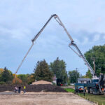 Construction scene with two large concrete pump trucks forming arches as they pour cement on a site. Workers are visible with soil piles and trees nearby, under a cloudy sky.