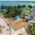 Aerial view of a lakeside park with a statue of a lumberjack and blue ox. Nearby, a cement truck and crane are working on path construction. Trees and parked cars line the area, with a lake in the background.