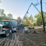 Concrete mixer trucks lined up on a dirt road in a forest clearing, unloading cement through a long boom. Workers in safety gear operate nearby. Autumn trees surround the site.