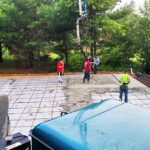 Workers pour concrete on a large grid-patterned foundation, surrounded by lush green trees. A blue truck is visible in the foreground.